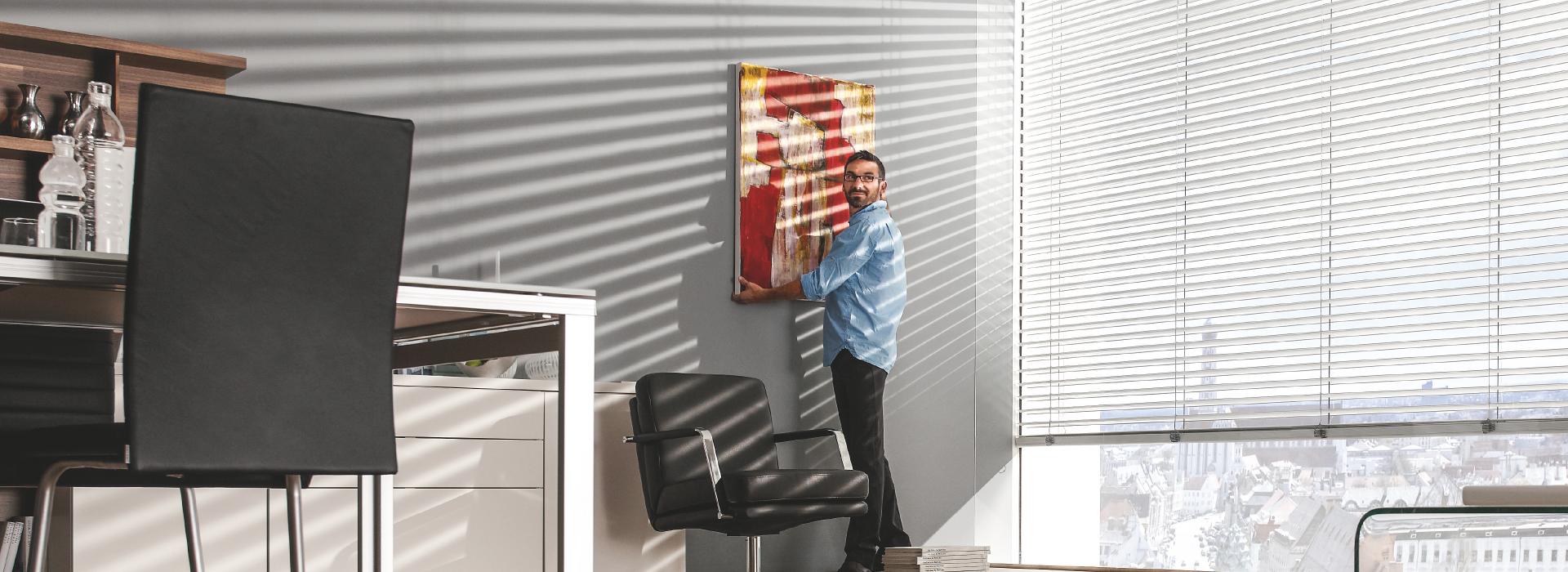Mann hängt Gemälde in Büro auf, Raffstoren sind gekippt