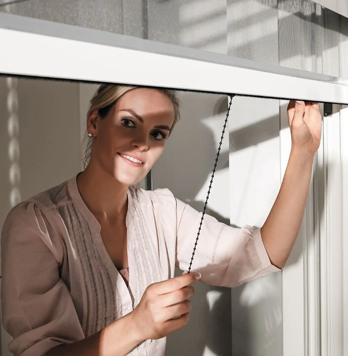 Frau vor Fenster zieht Insektenschutzgitter runter und freut sich auf ein Leben ohne lästige Fluginsekten im Haus