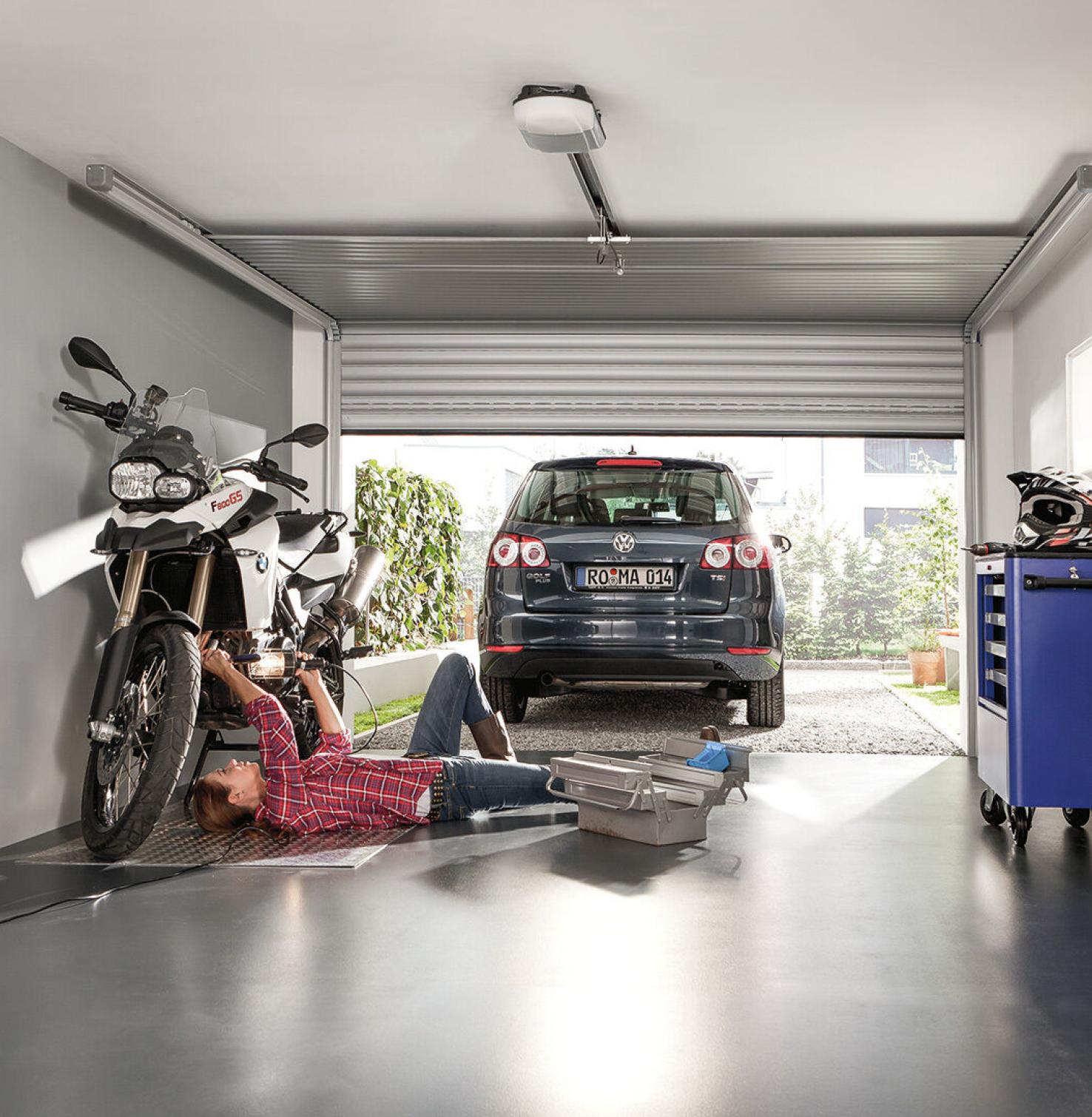 Frau schraubt liegend an Motorrad in Garage, davor steht ein Auto